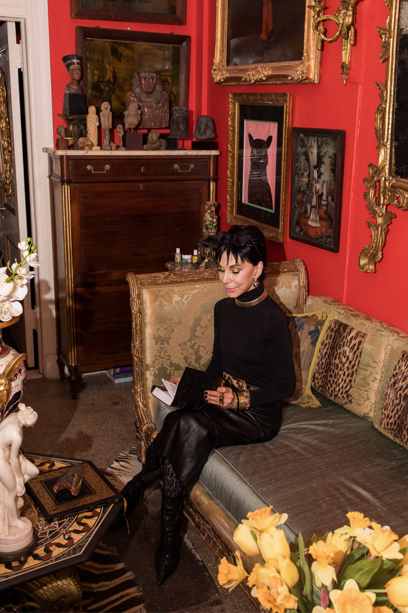 Cynthia is seated upon an 18th-century Italian daybed upholstered in luminous sage silk and framed by vintage ocelot accents and an 18th century Aubusson tapestry pillow. Before her rests an 18th-century pietra dura table adorned with classical Greci