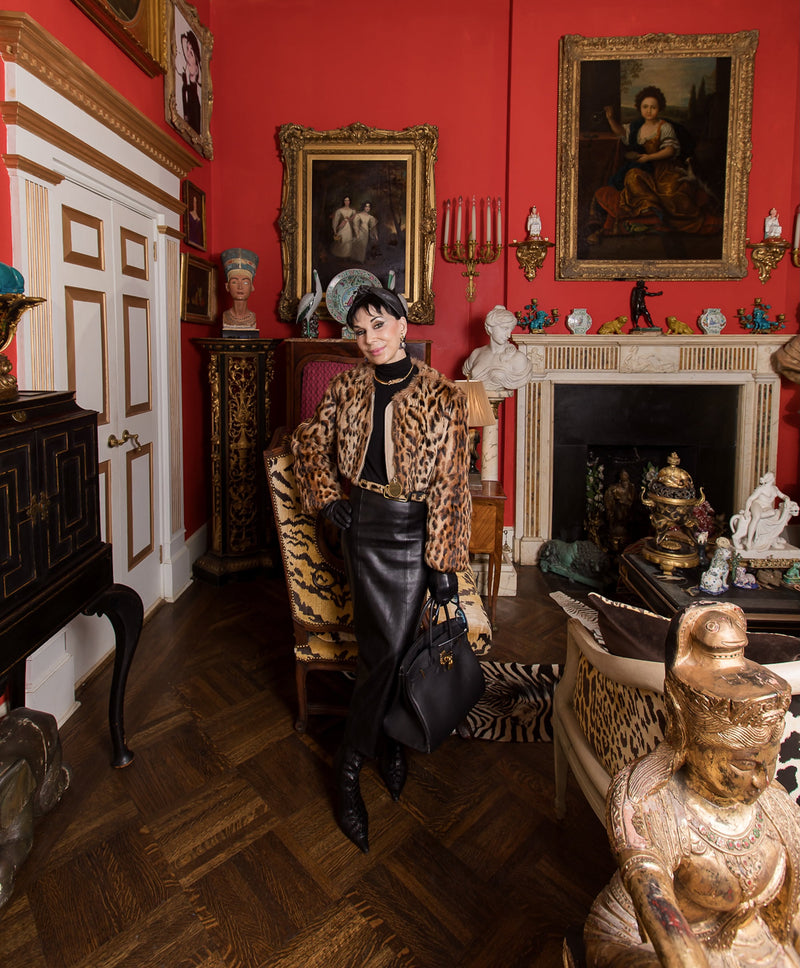 Cynthia appears in a Givenchy leopard print jacket, paired with a disciplined black silhouette and archival accessories. A salon-style arrangement of European paintings unfolds across crimson walls, anchored by an imported 18th-century Regency marble